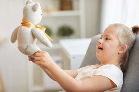 Down syndrome girl sitting on armchair and playing with toy bear at homeの写真素材