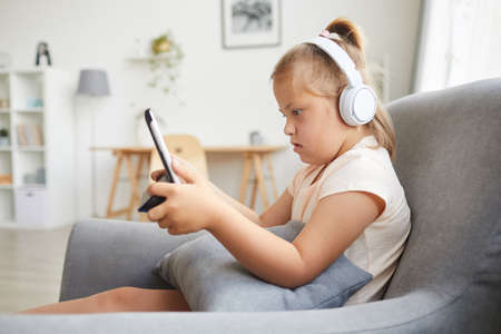 Down syndrome girl in wireless headphones using digital tablet while resting on armchair in the living roomの写真素材