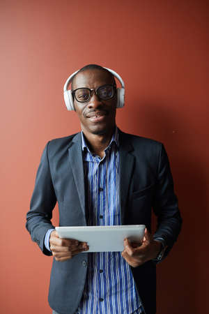 Portrait of African young man in eyeglasses and wireless headphones smiling at camera while using...