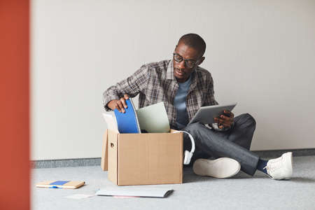 African young man sitting on the floor unpacking the box with documents and using digital tabletの写真素材