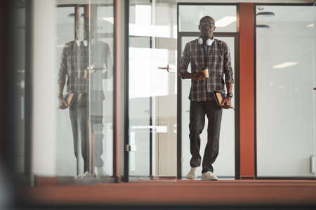 African young businessman in casual clothing walking along the office corridor and drinking coffeeの写真素材