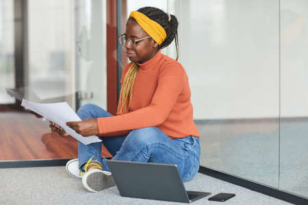 African businesswoman in casual clothing sitting on the floor near the laptop and examining...