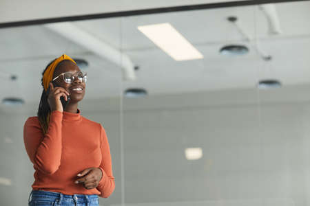 African young woman in eyeglasses talking on mobile phone while standing at officeの写真素材