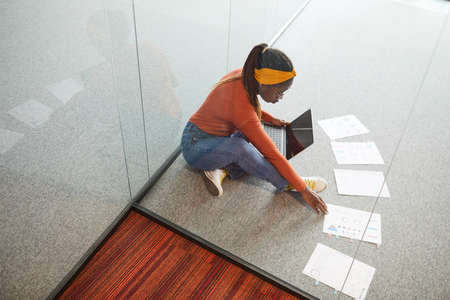 Young designer sitting on the floor and examining documents with charts using her digital tablet at officeの写真素材