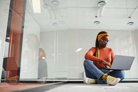 Designer in casual clothing sitting on the floor and working online using her laptop computer at...