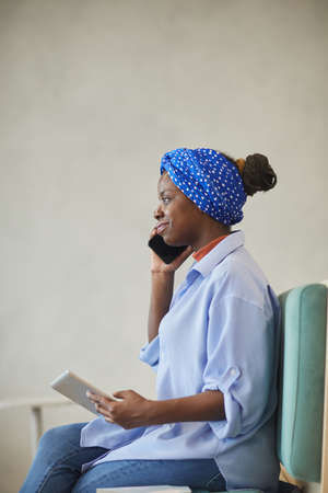 African young businesswoman talking on the mobile phone and using digital tablet while sitting on...