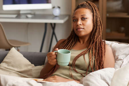 Portrait of young woman with beautiful hairstyle looking at camera while resting on sofa and...