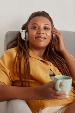 Portrait of African woman in headphones smiling at camera while resting on armchair with cup of...