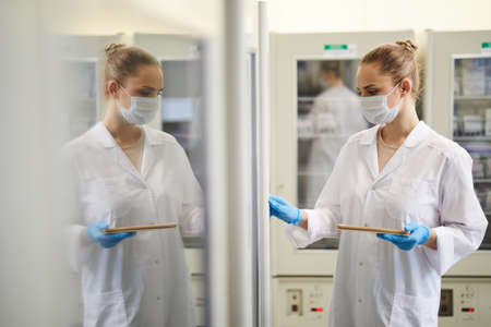 Female doctor in mask and in white coat using digital tablet while working at the laboratory