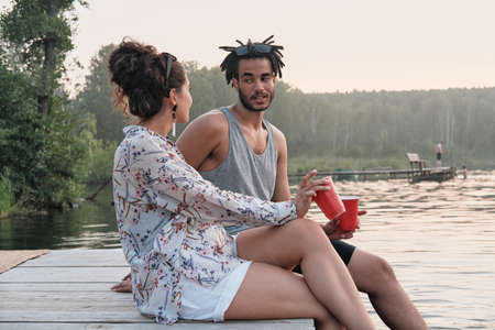 Young couple sitting on a pier with cocktails and talking to each other during summer day