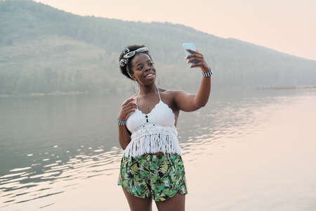 African young woman standing against the mountain and the lake and making selfie on her mobile phoneの写真素材