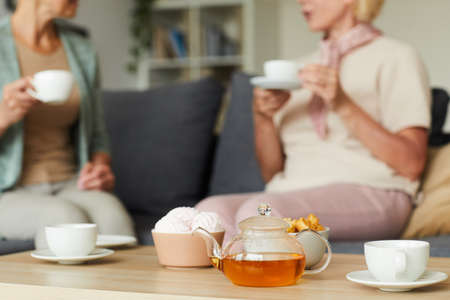 Close-up of teapot with tea with cups on the table and two women talking to each other on the sofa in the backgroundの写真素材