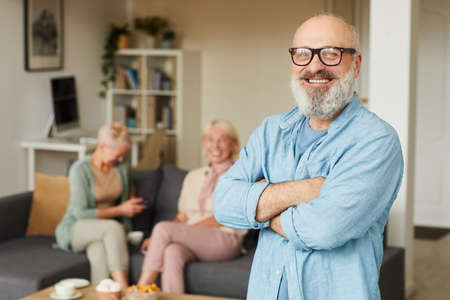 Portrait of mature bearded man in eyeglasses standing with arms crossed and smiling at camera with two women in the background in the roomの写真素材