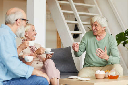 Senior man talking to his friends while they sitting on sofa and drinking tea in the roomの写真素材