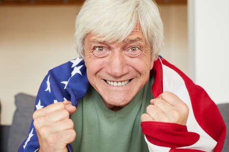 Portrait of excited senior man with white hair with American flag on his back watching football at homeの写真素材