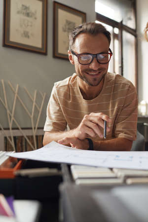 Mature designer in eyeglasses sitting at the table looking at blueprint and smiling he working at officeの写真素材
