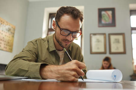 Mature designer in eyeglasses sitting at the table and drawing the blueprint of new project at...