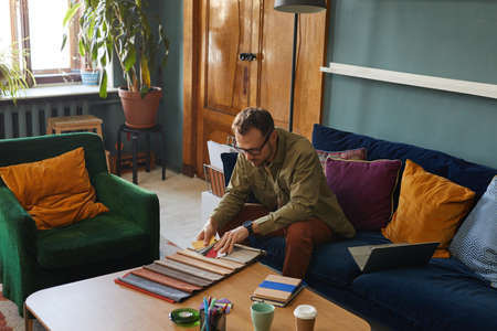 Mature designer in eyeglasses sitting on sofa in front of the table and working with fabric samples at modern officeの写真素材