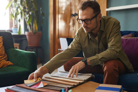 Mature designer in eyeglasses sitting at the table and choosing the color of fabric he working with...
