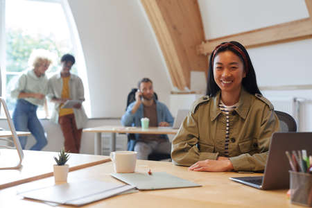 Portrait of Asian young woman smiling at camera while working at her workplace with other people in...
