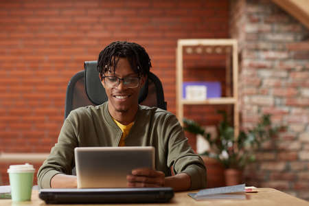 African young businessman sitting at the table and working online on digital tablet at office he...