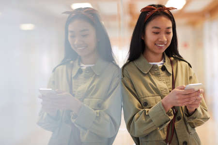 Asian young woman in casual clothing smiling while typing a message on her mobile phone