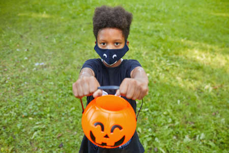 African boy in Halloween costume holding pumpkin basket and asking for treats while standing outdoorsの写真素材
