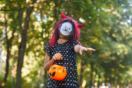 Spooky little girl in Halloween costume with pumpkin bag playing outdoors