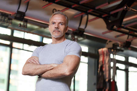 Portrait of mature muscular instructor standing with his arms crossed and looking at camera in the gymの写真素材