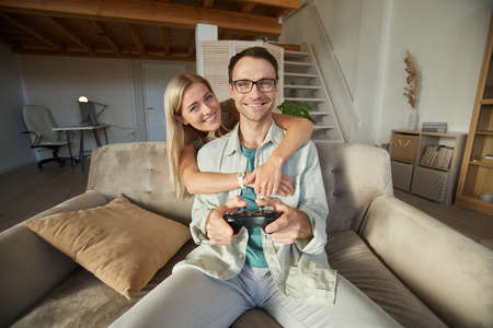 Portrait of young happy woman embracing her boyfriend while he playing in computer game they sitting on sofa in the living roomの写真素材