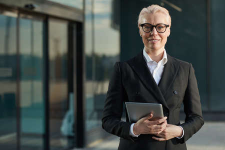 Portrait of mature businesswoman in eyeglasses and in black elegant suit holding digital tablet and...