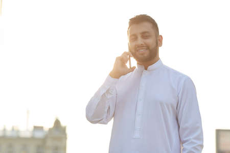 Portrait of young businessman in traditional costume smiling during conversation on mobile phone...