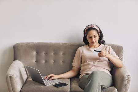 Young woman looking at number of her credit card while sitting on sofa and using laptop for online shoppingの写真素材