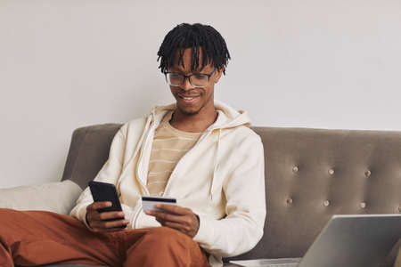 African happy man sitting on sofa and using his mobile phone to paying bills online with credit cardの写真素材