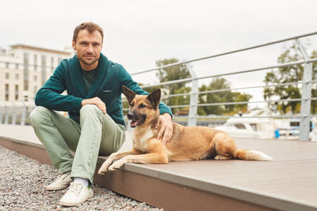 Portrait of young man looking at camera while sitting with his dog on the street outdoorsの写真素材