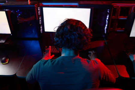 Rear view of young hacker sitting at the table in front of computer monitor working with software in...
