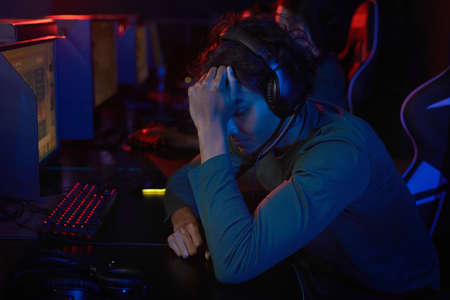Tired developer in headphones sitting at the table in front of computer and holding his head he has...