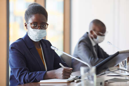 Portrait of African businesswoman in protective mask looking at camera while working at conference during pandemicの写真素材