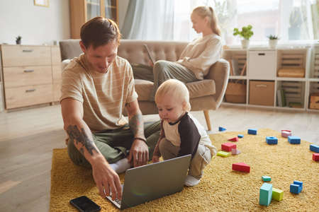 Young father sitting on the floor and watching cartoons on laptop together with his son with mother in the backgroundの写真素材