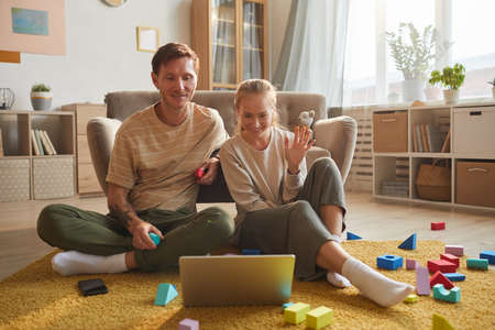 Young couple sitting on the floor smiling and waving looking at monitor of laptop they talking...