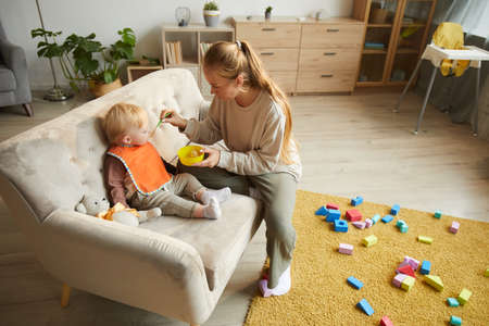 Young mother feeding her baby on the sofa in the living room