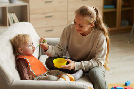 Smiling young mother feeding child with spoon during lunch time at home