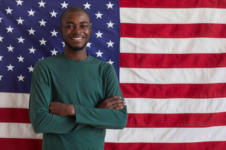 Portrait of young Afro-American man standing with arms crossed and smiling at camera against the American flagの写真素材