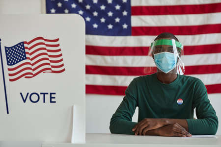 Portrait of African man in protective mask sitting at the table and working in voting at American polling stationの写真素材