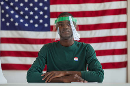 Portrait of Afro-American man wearing protective mask sitting at the table and voting for American...
