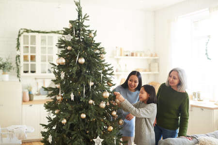 Little girl hanging Christmas balls on Christmas tree together with her elder sister and mother in...