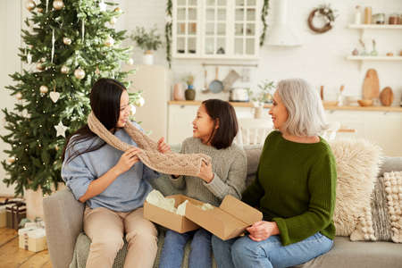 Mother and little sister sitting on sofa and giving warm scarf for elder daughter on Christmas in...