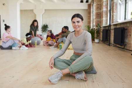 Portrait of young dancer sitting on the floor in dance studio and smiling at camera with other group in the backgroundの写真素材
