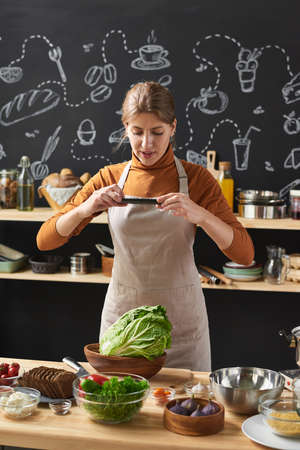 Young woman in apron making photo of fresh vegetables in bowl on her mobile phone standing in the...