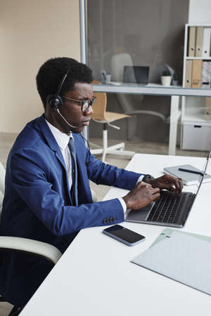 African businessman in headphones communicating online on laptop computer while working at the table...